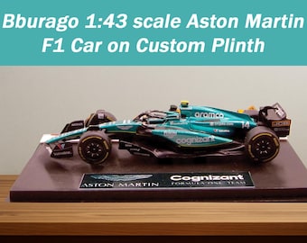 Puede incluir: Modelo a escala 1:43 de un coche Aston Martin F1, de color verde azulado y negro, sobre una base personalizada. La base es de color marrón oscuro y presenta los logotipos de Aston Martin y Cognizant. El texto "Bburago 1:43 scale Aston Martin F1 Car on Custom Plinth" está en la parte superior.
