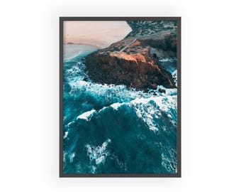 May include: An aerial view of a rocky coastline with crashing waves. The water is a deep blue with white foam. The rocks are brown and gray. The sky is a light pink and orange.