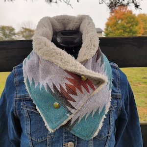 May include: A cozy, patterned neck warmer with a soft, cream-colored fleece lining. The scarf features a geometric design in shades of gray, teal, and rust, secured with two gold-toned buttons. It is worn over a denim jacket.