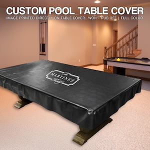May include: A black custom pool table cover with the name "MARTINEZ" in a white decorative frame. The cover is draped over a pool table with a dark wood base. The image also shows an air hockey table and a staircase.