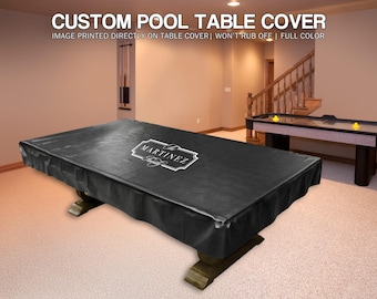 May include: A black custom pool table cover with the name "MARTINEZ" in a white decorative frame. The cover is draped over a pool table with a dark wood base. The image also shows an air hockey table and a staircase.