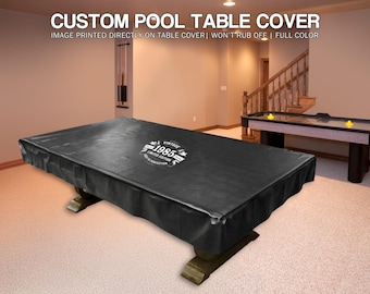 May include: A black custom pool table cover with white text that reads "VINTAGE 1985 LIMITED EDITION". The cover is on a pool table in a room with a second table and stairs.