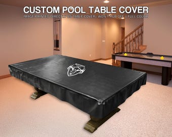 May include: A black custom pool table cover with white text that reads "CUSTOM POOL TABLE COVER". The cover is on a large rectangular pool table with a logo in the center. The image also shows an air hockey table.