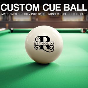 May include: A white cue ball with a custom design featuring the name "Reynolds" and the letter "R" in a decorative font. The ball is on a green pool table. Text at the top reads "CUSTOM CUE BALL".