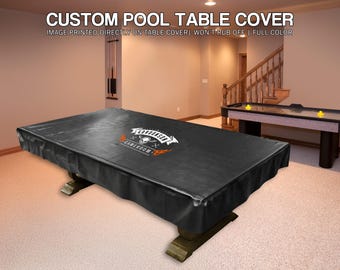 May include: A black custom pool table cover with the text "CUSTOM POOL TABLE COVER" and "GAMEROOM" printed on it. The cover is on a wooden pool table in a room with an air hockey table.