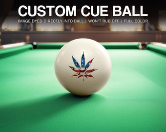 May include: A custom cue ball with a cannabis leaf design in red, white, and blue, resting on a green pool table. The text "CUSTOM CUE BALL" is at the top of the image.