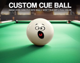 May include: A white custom cue ball with a surprised face graphic, resting on a green pool table. The text "CUSTOM CUE BALL" is at the top, with additional text indicating the image is dyed directly into the ball and won't rub off.