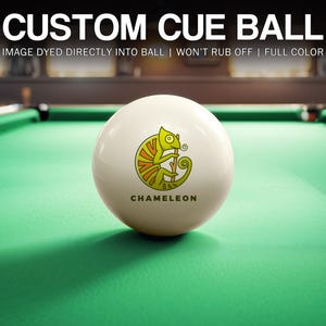 May include: A white cue ball with a chameleon graphic and the word "CHAMELEON" printed on it. The ball is on a green felt pool table.
