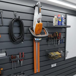 Peut inclure: Un système d'organisation de garage avec un taille-haie STIHL noir et orange, une rallonge électrique noire enroulée et divers outils à main. Une étagère contient des bombes aérosols et un panier en fil de fer contient des gants et des lunettes de sécurité. L'étiquette murale indique "OUTILS DE JARDIN."
