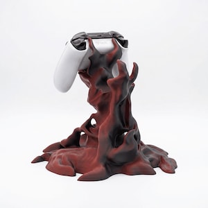 May include: A white Xbox controller sits on a decorative stand. The stand has a dark red and black abstract design, with a textured, organic appearance. The controller has black accents.