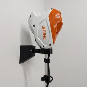 May include: A white and orange STIHL power tool head mounted on a black bracket and pole. The STIHL logo is visible on the side of the tool. The tool is mounted on a white wall.