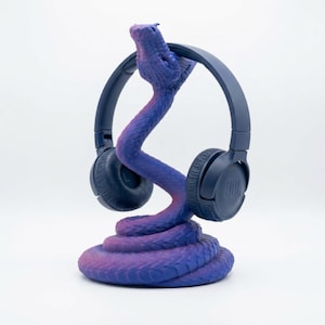 Op de afbeelding: Een marineblauwe JBL-koptelefoon rust op een paars en blauw slangvormig hoofdtelefoonstandaard. De standaard is aan de basis opgerold en heeft een textuur met schubben. De kop van de slang ondersteunt de koptelefoon.