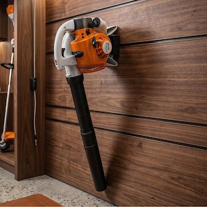 Puede incluir: Un soplador de hojas de gasolina STIHL BG 86 naranja y blanco montado en una pared de madera. El soplador tiene una boquilla negra y un mango blanco. Un cortabordes es visible en el fondo.