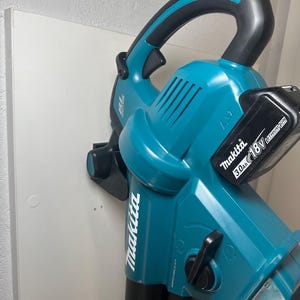 Op de afbeelding: Een draadloze Makita bladblazer in teal en zwart. De blazer heeft een gebogen handvat en een batterij. Het Makita-logo is zichtbaar aan de zijkant. De batterij heeft de tekst "3.0Ah 18V Lithium Ion".
