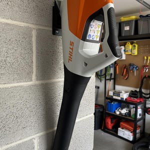 Puede incluir: Un soplador de hojas inalámbrico de la marca Stihl montado en una pared de bloques de hormigón gris. El dispositivo cuenta con una carcasa blanca y naranja con un mango negro y una boquilla larga negra. Se ve una etiqueta de advertencia con iconos.