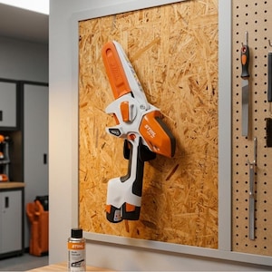 Könnte beinhalten: Eine weiß-orangefarbene Stihl GTA 26 Akku-Gehölzschneider, die an einer Lochwand hängt. Das Werkzeug verfügt über eine kompakte Führungsschiene, einen schwarzen Griff und einen Akku an der Basis. Der Markenname Stihl ist auf dem Gehäuse aufgedruckt.