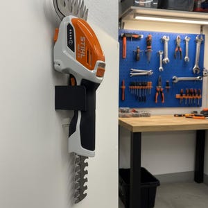 Op de afbeelding: Een oranje en witte STIHL HBA 25 snoerloze heggenschaar gemonteerd op een witte muur. De trimmer heeft een zwarte handgreep en een zilveren mes. De afbeelding toont een werkplaats met gereedschap en een werkbank op de achtergrond.