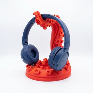 May include: Navy blue JBL headphones resting on a bright orange, tentacle-shaped headphone stand. The stand has a textured surface and is designed to hold the headphones securely. The JBL logo is visible on the ear cup.