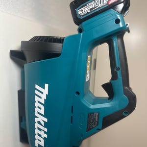 Puede incluir: Un soplador de hojas inalámbrico Makita, turquesa y negro, con una batería de 40V adjunta. El logotipo de Makita se muestra en blanco. El soplador tiene un asa y un gatillo.