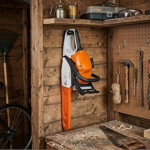 Op de afbeelding: Een oranje en witte kettingzaag is aan een houten muur bevestigd. De kettingzaag heeft een zwarte handgreep en een oranje beschermkap. De achtergrond omvat een werkbank, gereedschap en een plank met diverse items.