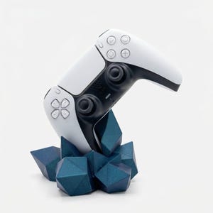 Peut inclure: Une manette de jeu blanche et noire est présentée sur un support géométrique bleu. La manette a un design épuré avec des boutons et des joysticks. Le support est composé de plusieurs formes bleues angulaires.