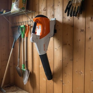 Op de afbeelding: Een wit en oranje Stihl bladblazer gemonteerd aan een houten wand. Het handbediende elektrische gereedschap is voorzien van een zwarte blaasmond, een zwarte handgreep en een opvallende oranje motorbehuizing met de merknaam Stihl in zwarte letters.