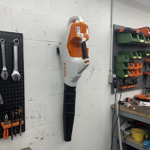 Può includere: Un soffiatore per foglie Stihl bianco e arancione montato verticalmente su una parete in blocchi di cemento bianchi. L'utensile presenta un'impugnatura nera, un lungo beccuccio nero e il marchio Stihl stampato in arancione sul corpo bianco.