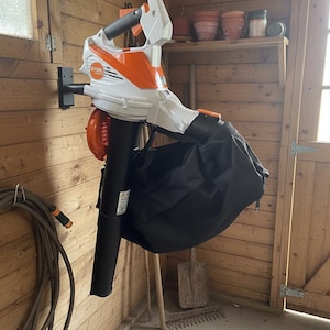 Peut inclure: Un souffleur de feuilles STIHL blanc et orange avec un sac de collecte noir est monté sur un mur d'abri de jardin en bois. Le souffleur est conçu pour les travaux de jardinage et possède une buse et une poignée noires.