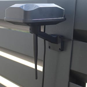 Op de afbeelding: Een zwart, rechthoekig apparaat met een gebogen bovenkant en twee antennes, gemonteerd op een donkergrijze metalen oppervlakte. Het apparaat is bevestigd aan een beugel en lijkt een antenne of ontvanger te zijn.