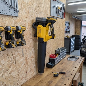 May include: A yellow and black DeWalt cordless leaf blower mounted on a wooden wall, positioned next to two yellow and black cordless impact drivers. The blower features a long black nozzle and a battery pack attached to the handle area.