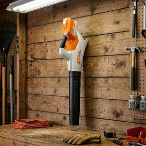 Peut inclure: Un souffleur de feuilles STIHL orange et blanc suspendu à un mur en bois. Le souffleur a une poignée et une buse noires. Un établi avec des outils, des gants et une rallonge orange est au premier plan. Le mot "STIHL" est imprimé sur le côté.