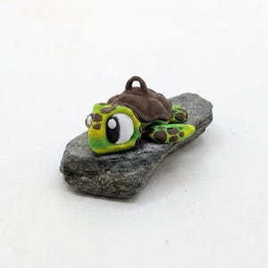 May include: A 3D-printed turtle figurine with a brown shell, green head, and yellow and green flippers. The turtle has large white eyes and is resting on a grey stone. The figurine has a loop on top of the shell.