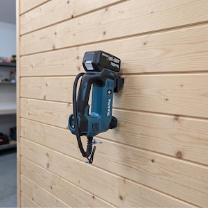 Könnte beinhalten: Ein türkisfarbenes und schwarzes Makita Akku-Elektrowerkzeug mit angebrachtem 18V-Akkupack, montiert an einer Wand aus hellen, horizontalen Holzplanken. Das Werkzeug hat einen schwarzen Griff, ein Stromkabel und den weißen Makita-Schriftzug.