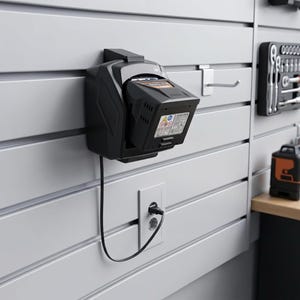 Puede incluir: Un cargador de batería negro montado en una pared blanca. El cargador tiene una batería insertada y está enchufado a una toma de corriente. La pared tiene ranuras horizontales. Una caja de herramientas y otras herramientas son visibles en el fondo.