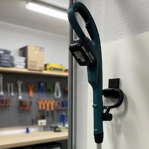 Op de afbeelding: Een turquoise en zwarte draadloze grastrimmer gemonteerd op een witte muur. De trimmer heeft een zwart batterijpakket en een zwarte handgreep. Een zwarte muurbevestiging houdt de trimmer vast. De achtergrond toont een werkplaats met gereedschap.