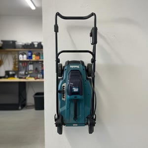 May include: A teal and black Makita cordless lawn mower hanging on a white wall. The mower has a black handle and wheels. The background shows a garage with tools and equipment.