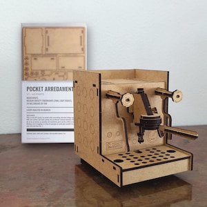 Könnte beinhalten: Ein lasergeschnittenes Holzmodell einer Espressomaschine aus mitteldichter Faserplatte. Das Set enthält einen Bogen mit 40 Teilen und eine Schachtel mit der Aufschrift POCKET ARREDAMENTO V1. Das zusammengebaute Modell zeigt einen Siebträger und Knöpfe.
