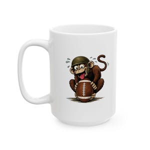 May include: White ceramic mug featuring a cartoon monkey wearing a helmet and holding a football. The monkey has a surprised expression with its tongue sticking out. The mug is a standard size and has a large handle.
