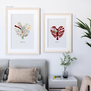 K&ouml;nnte beinhalten: Zwei gerahmte Kunstdrucke. Einer zeigt eine Herzform aus bunten Blumen, der andere ein rotes Rippenherz mit Blumen. Beide sind in hellen Holzrahmen und haben unten Text. Die Drucke werden &uuml;ber einem Bett ausgestellt.