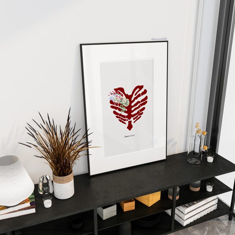 K&ouml;nnte beinhalten: Gerahmter Kunstdruck mit einem roten Brustkorb-Herz-Design und einem Blumenarrangement in der Mitte. Das Kunstwerk wird in einem schwarzen Rahmen vor einem wei&szlig;en Hintergrund pr&auml;sentiert. Der Text "Genesis 2:23-25" befindet sich unten.