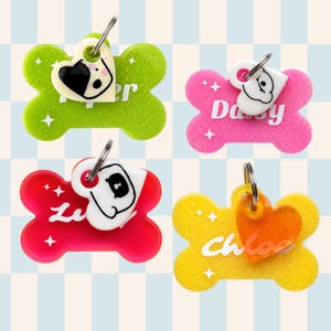 May include: Four colourful dog tags in the shape of a bone. Each tag has a different colour: green, pink, red, and yellow. Each tag has a different name and a charm with a dog or heart design. The tags are attached to a silver ring.