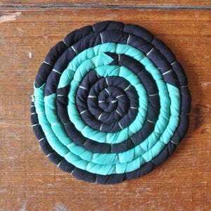 Pode incluir: Um descanso de panela redondo, com um padrão espiral em tons de verde-azulado e preto. O descanso de panela é feito de tiras de tecido bem tecidas, criando uma superfície texturizada. Está sobre uma superfície de madeira rústica.