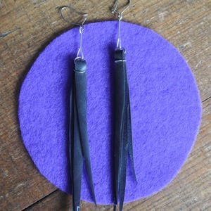 Op de afbeelding: Een paar zwarte leren kwast oorbellen met zilverkleurige oorhaken. De oorbellen worden getoond op een paarse vilten cirkel, tegen een houten ondergrond. De kwasten zijn lang en slank, met een puntig uiteinde.
