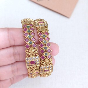 May include: Two gold bangles with detailed patterns. Each bangle has a repeating diamond pattern of red and green gemstones, with white gemstones and rectangular red stones. The bangles have an ornate, detailed appearance, suitable for special occasions.