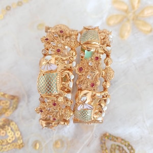 Peut inclure: Deux bracelets dorés ornés avec des motifs floraux et d'éléphants complexes. Les bracelets présentent des pierres précieuses rouges et transparentes, et des sections d'émail vert menthe et blanc. Les bijoux sont exposés sur une surface blanche texturée.