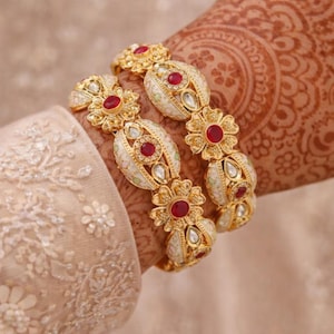 May include: Ornate gold bangles with red gemstone embellishments. The bracelets showcase floral and oval patterns with white enamel. The jewellery is worn on a hand adorned with henna, and a cream-coloured sleeve is visible.