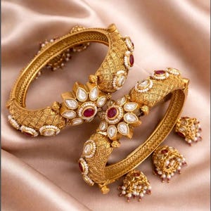 May include: Gold-toned jewelry set featuring two bangles and two pairs of earrings. The bangles and earrings are adorned with red and white gemstones, and small white pearls. The jewelry has an intricate, textured design.