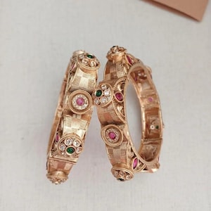 Könnte beinhalten: Zwei goldfarbene Armreifen mit aufwendigen Designs. Jeder Armreif weist eine Reihe von kreisförmigen und rechteckigen Mustern auf, die mit roten, grünen und klaren Edelsteinen verziert sind. Die Armreifen haben eine strukturierte Oberfläche und einen Vintage-Look.