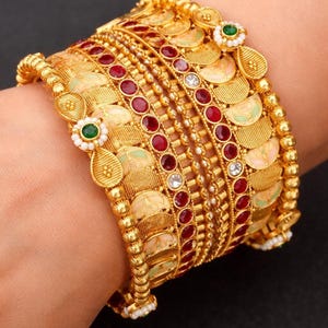 May include: A stack of gold bangles with intricate designs. The bangles feature red and white gemstones, along with green accents. The jewelry is ornate and detailed, with a mix of textures and patterns.