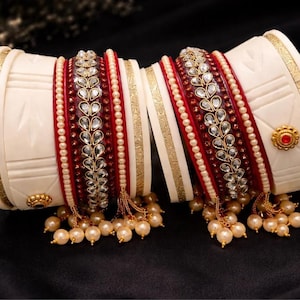 May include: Ivory and red bangles with gold accents and pearl embellishments. The bangles feature intricate designs with clear gemstones and a dangling pearl fringe. The jewelry is displayed on a black surface.